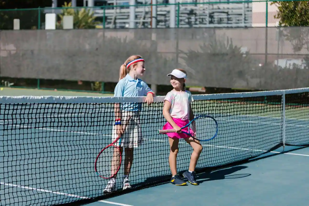Deux jeunes joueuses de tennis avec leurs raquettes, au bord du filet sur un court extérieur.