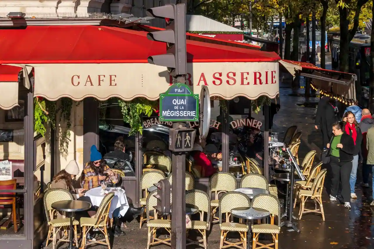 Terrasse d'un café-brasserie parisien à l'auvent rouge, avec tables rondes et chaises en rotin sur le trottoir, illustrant l'ambiance d'une brasserie de quartier.