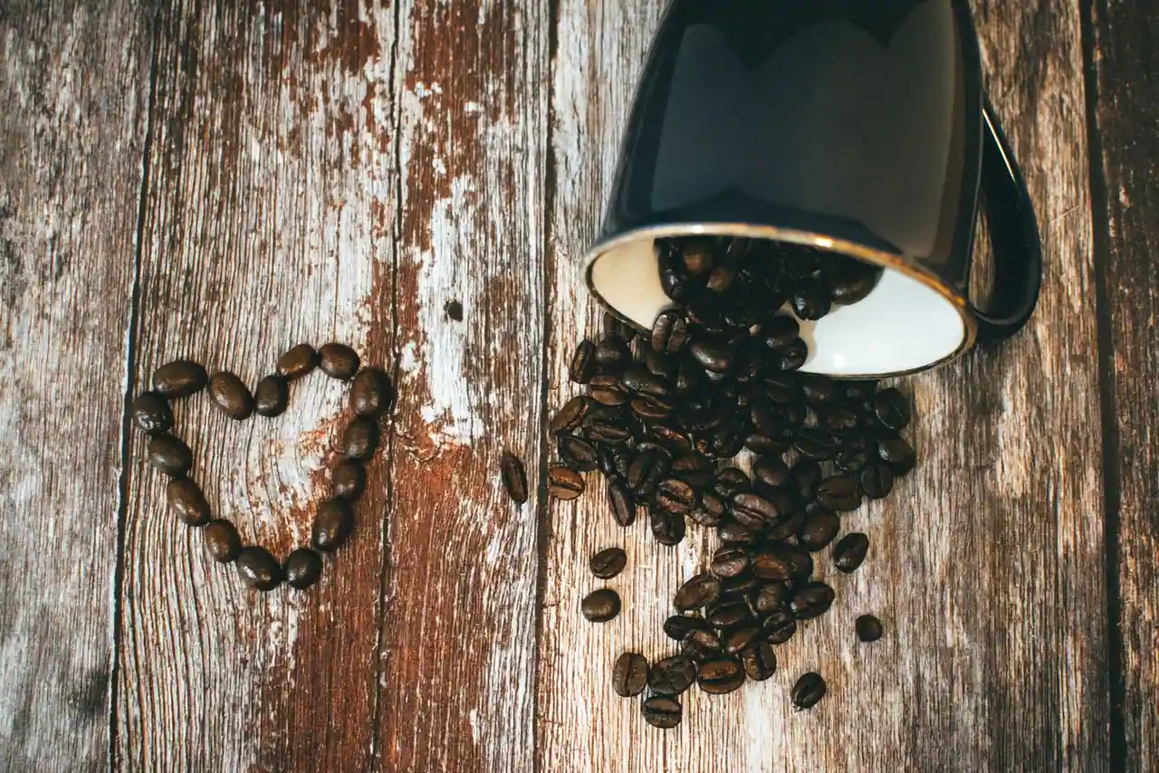 Mug noir renversé laissant échapper des grains de café torréfiés sur une table en bois, quelques grains formant un cœur.