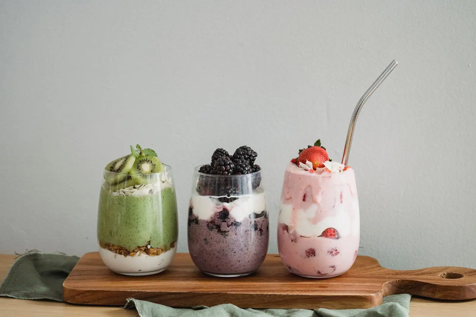 Trois verrines de fruits frais, yaourt et granola alignées sur une planche en bois clair.