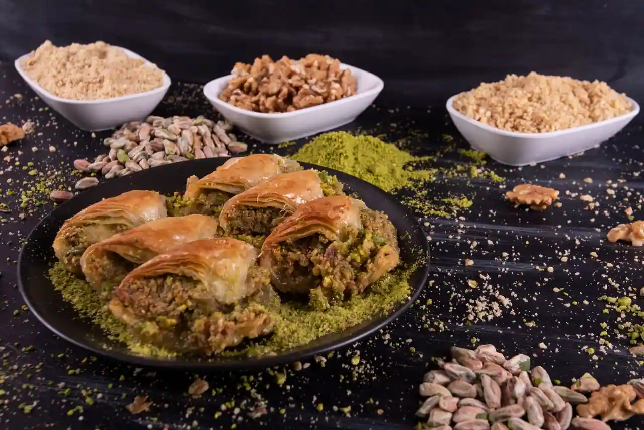 Parts de baklava garnies de pistache concassée sur une assiette sombre, entourées de fruits secs