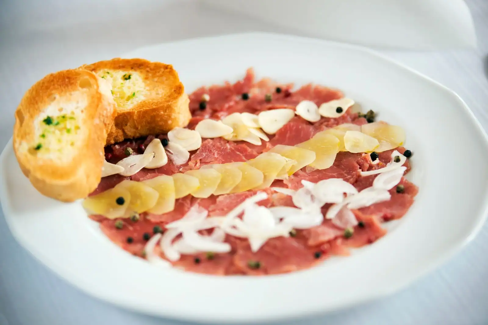 Carpaccio finement dressé sur une assiette blanche, accompagné de copeaux et de toasts dorés.