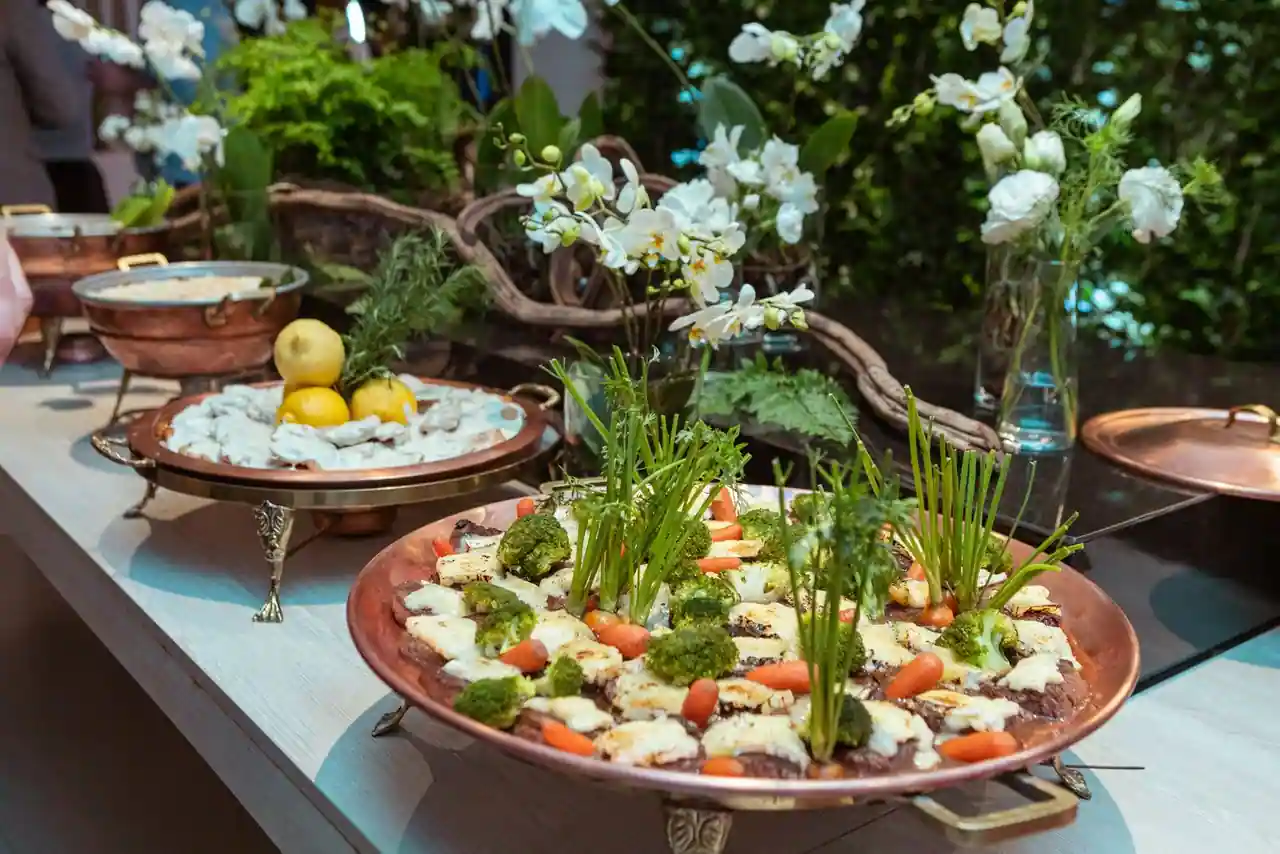 Grand plat en cuivre garni de légumes décorés et d'herbes sur une table de buffet fleurie.