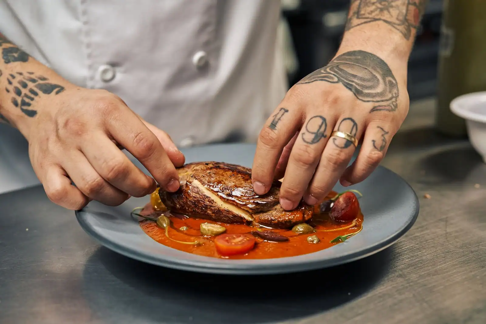 Mains d'un chef dressant un plat de viande nappé de sauce avec des tomates cerises sur une assiette.