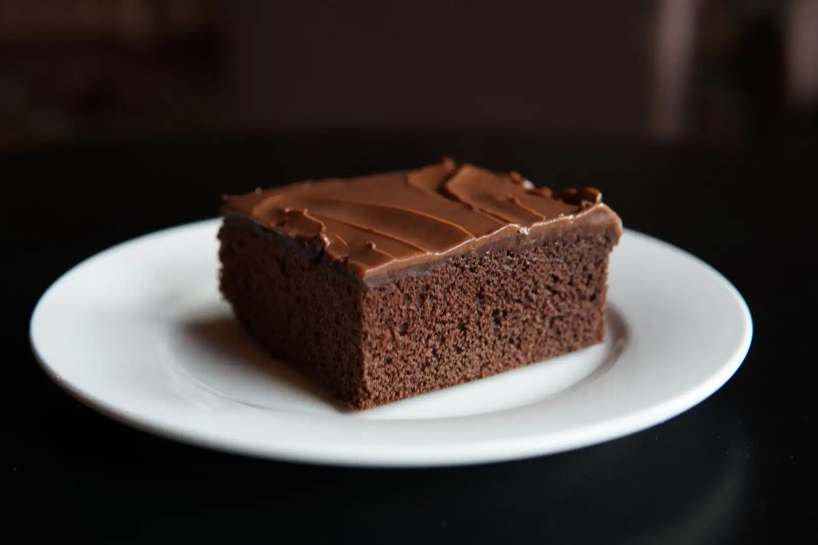 Part carrée de gâteau au chocolat nappée de glaçage au chocolat, posée sur une assiette blanche.