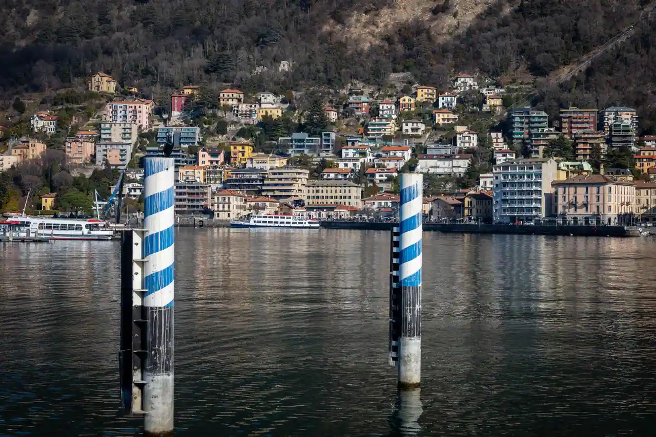 Rive du lac de Côme avec ses maisons étagées sur la colline et un ferry sur l'eau, deux pieux d'amarrage au premier plan.