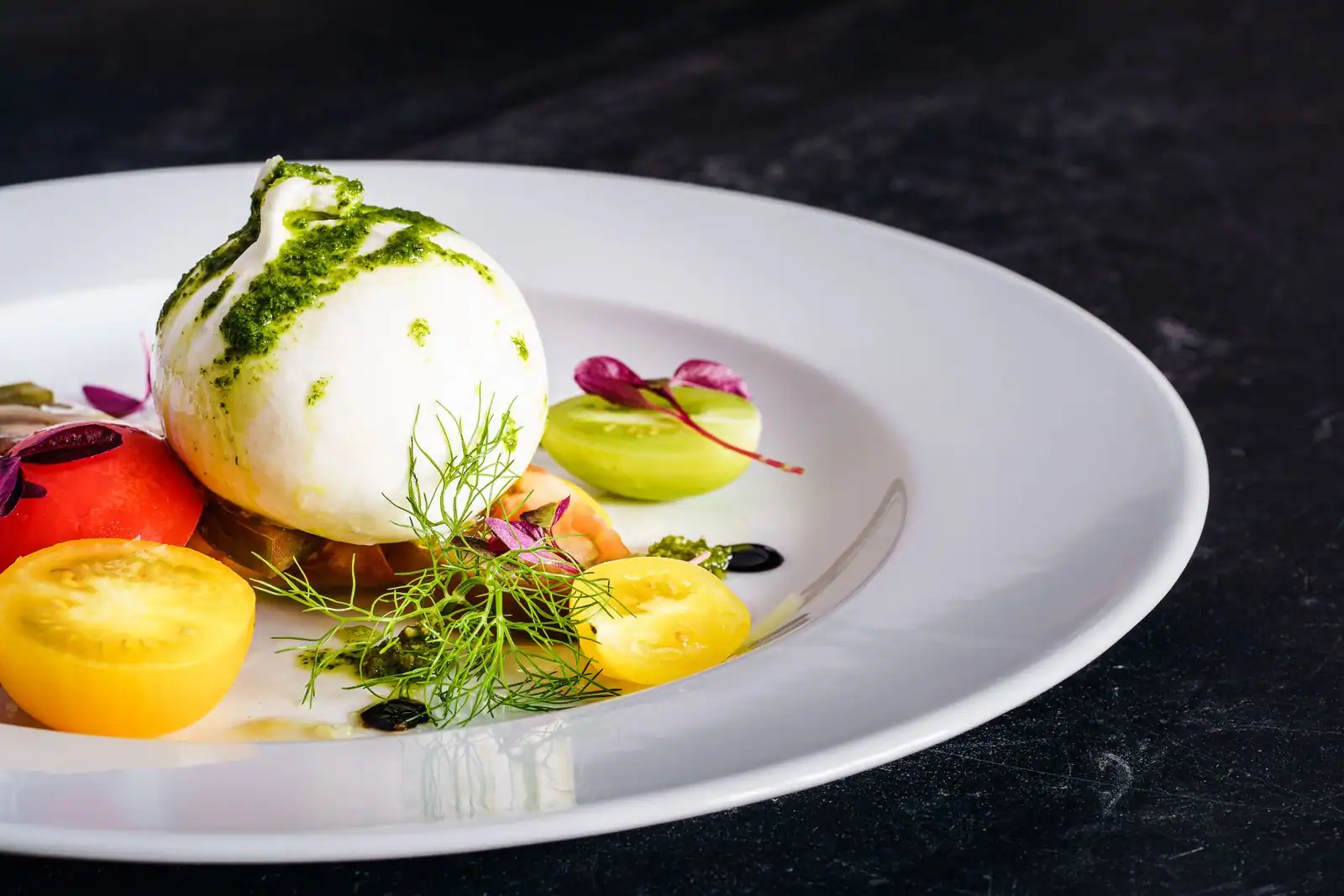 Entrée de burrata crémeuse sur des tomates colorées et un filet de pesto, dressée dans une assiette