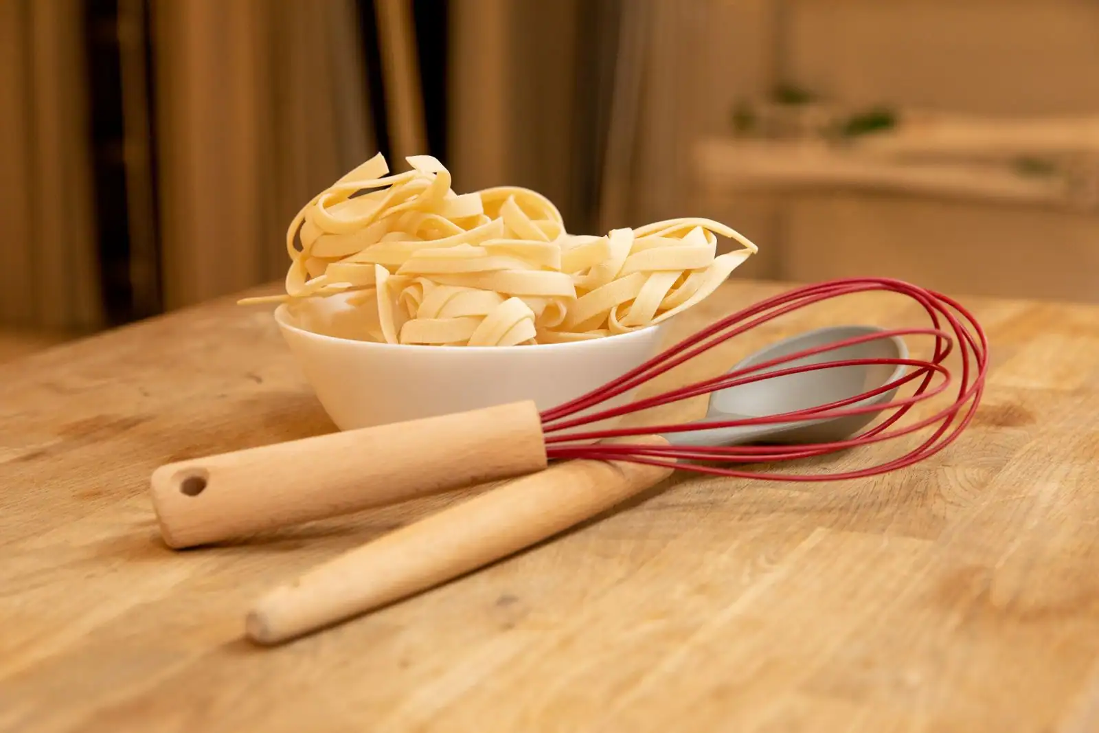 Un fouet rouge et un ustensile à manche en bois posés sur une table, à côté d'un bol de pâtes fraîches.