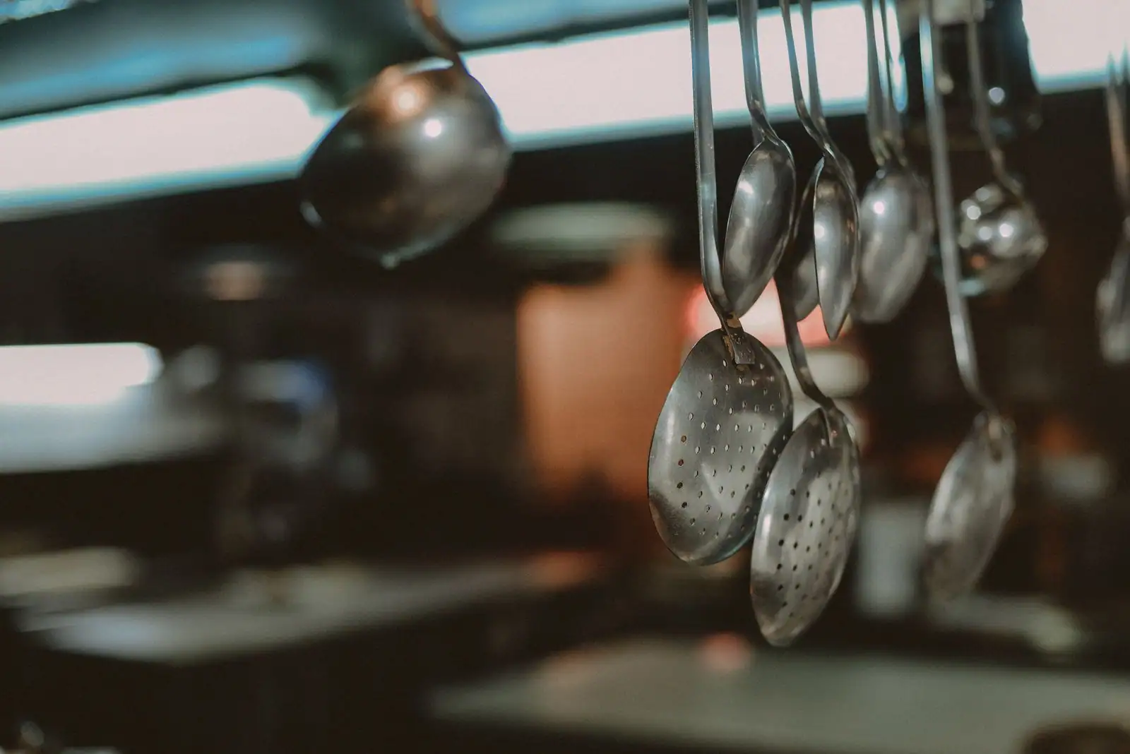 Louches et écumoires en métal suspendues à une barre dans une cuisine, sur un arrière-plan flou.