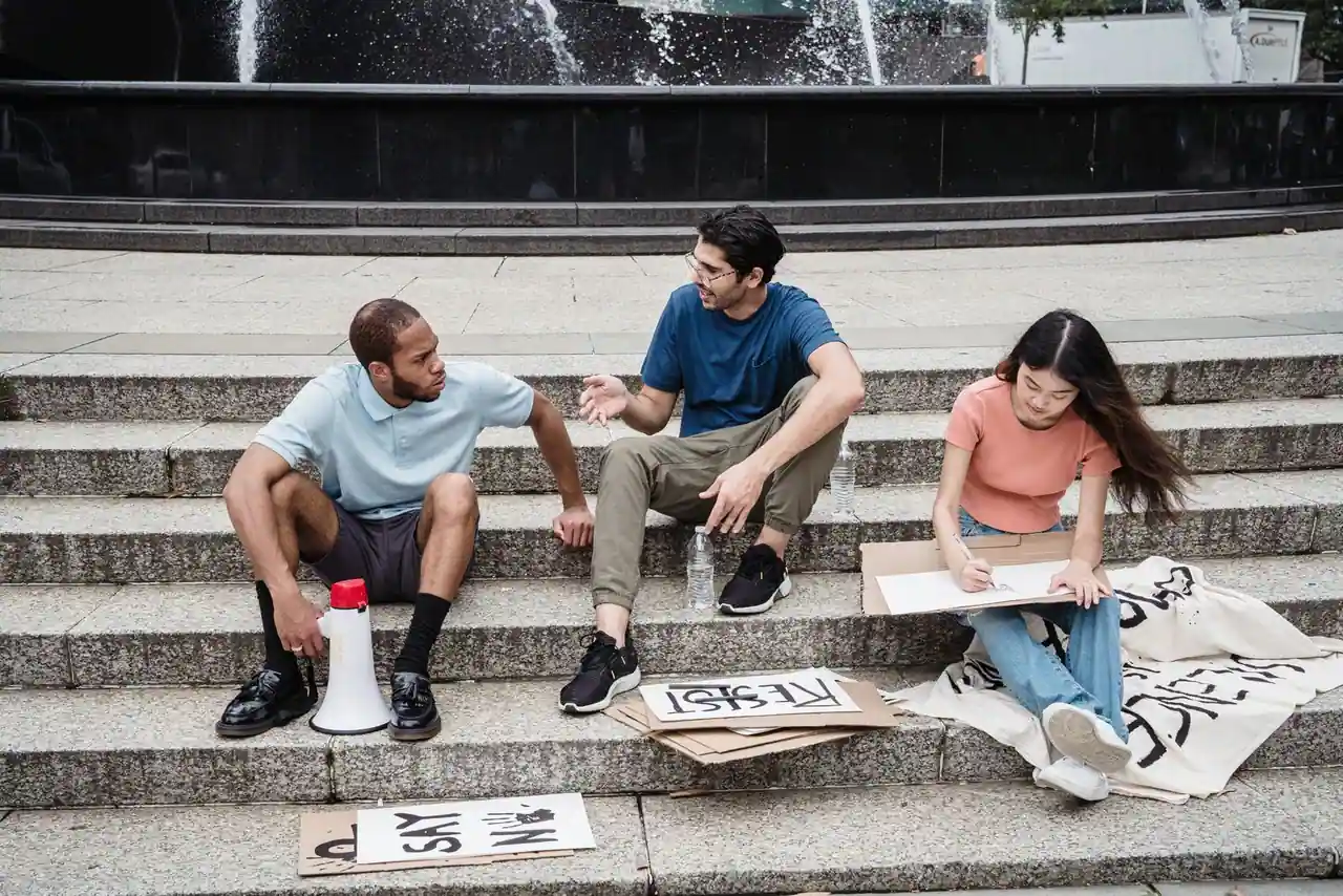Trois jeunes assis sur des marches dans un espace public, avec un porte-voix et des pancartes, illustrant l'engagement et le débat de société.