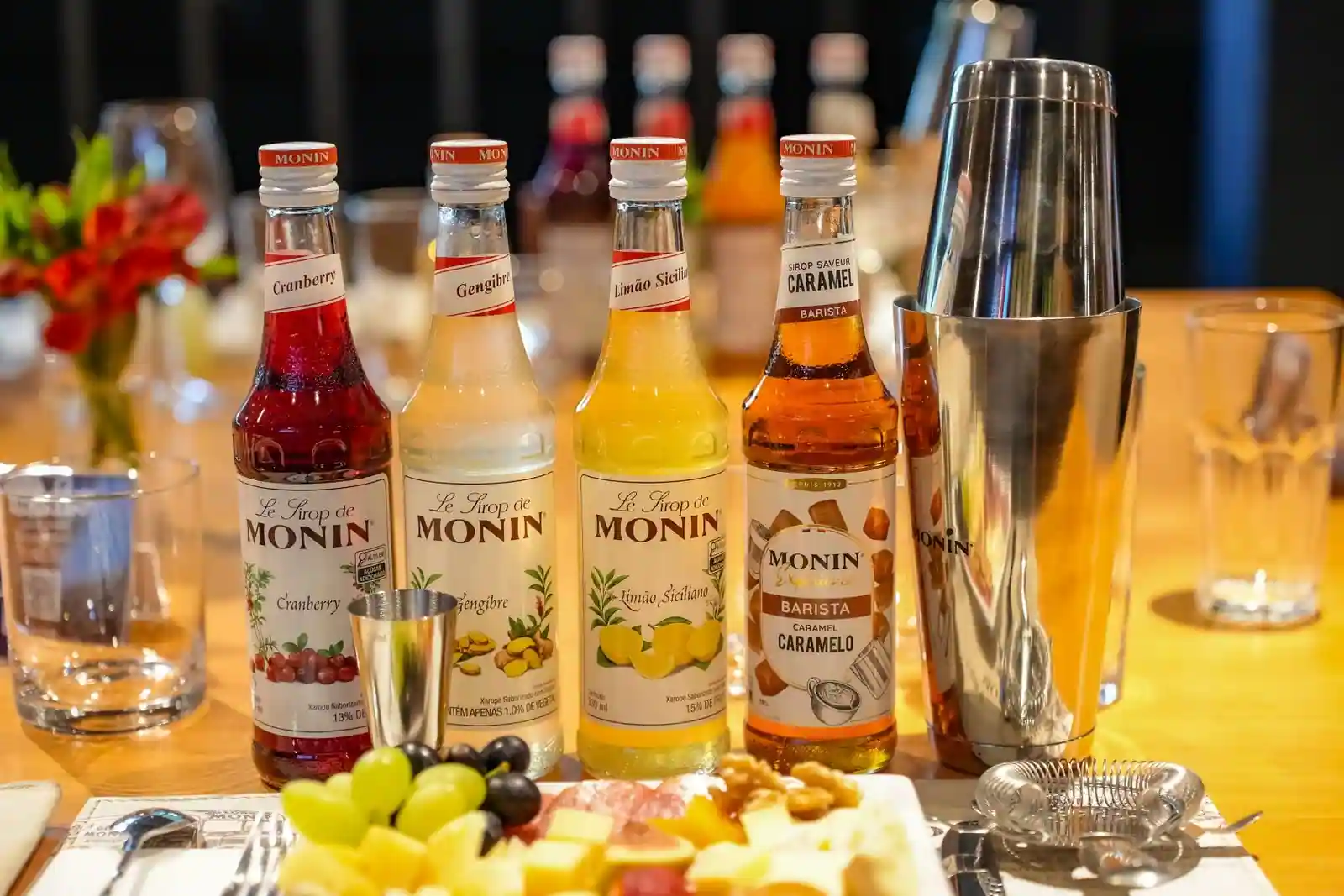 Bouteilles de sirops, shaker à cocktails, verres et doseur disposés sur une table en bois.