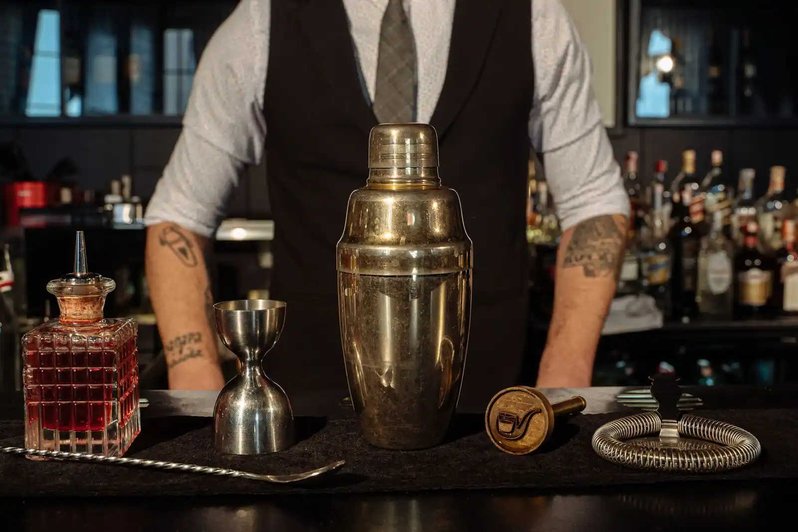 Barman en gilet derrière un comptoir de bar, avec shaker, doseur, cuillère à mélange et bouteilles en fond