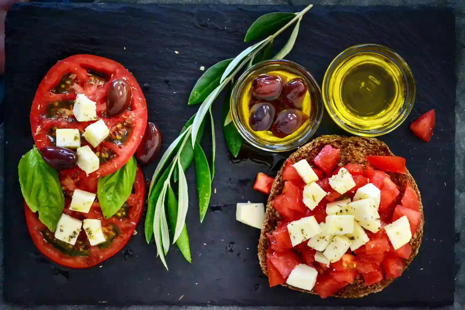 Ardoise garnie de tomates, de cubes de fromage blanc, d'olives noires, d'huile d'olive et d'un rameau d'olivier