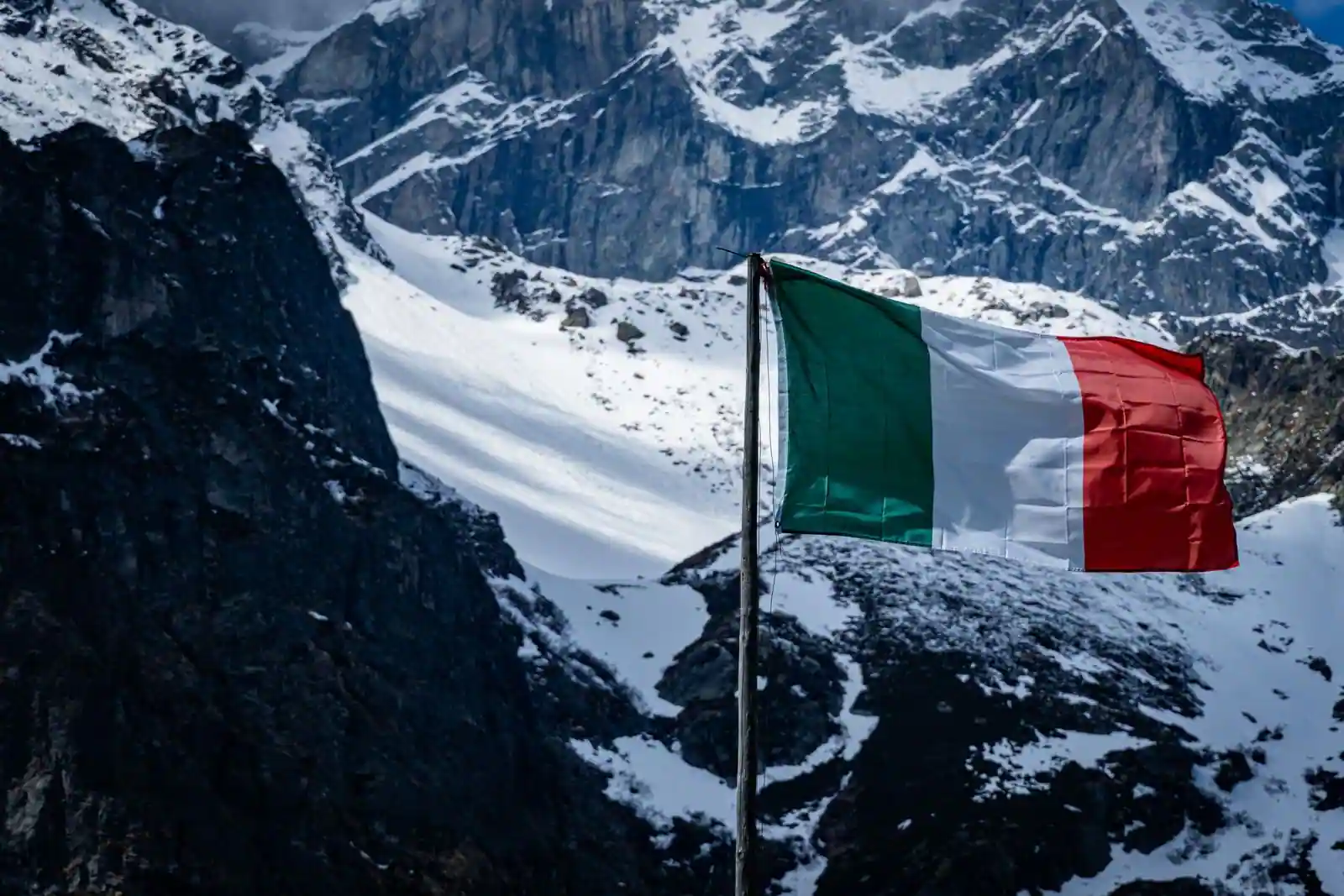 Drapeau italien vert, blanc et rouge flottant au vent devant un massif montagneux enneigé