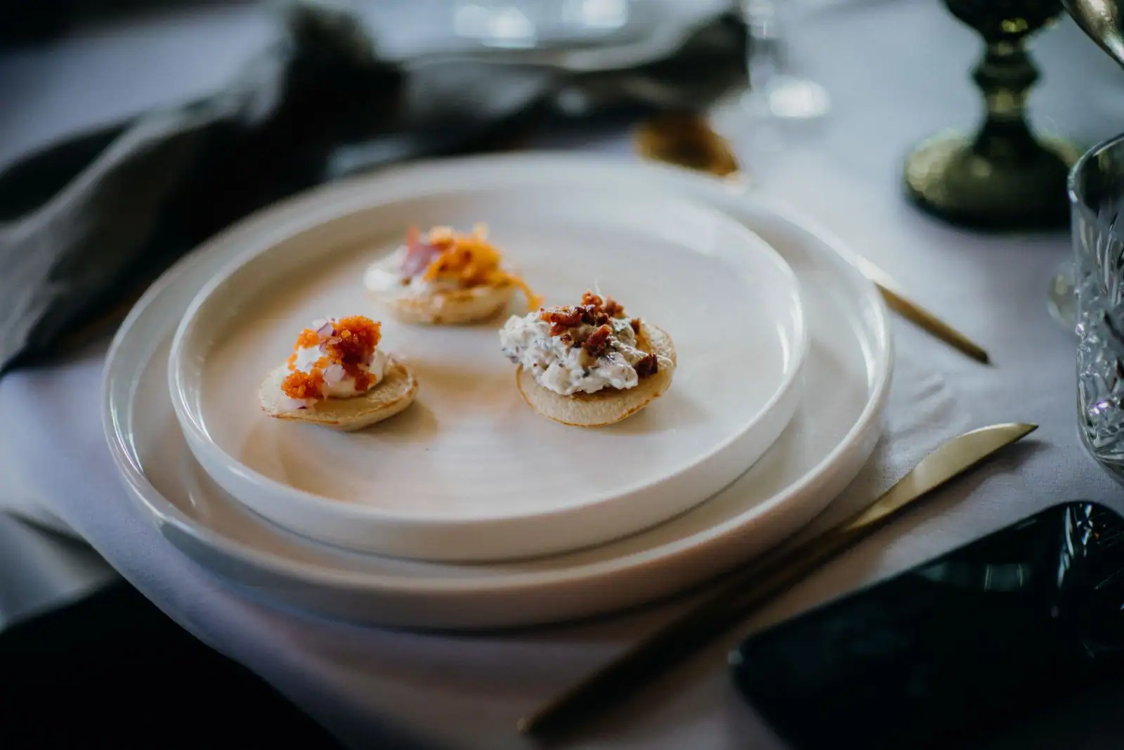 Petites entrées façon blinis garnis de fromage frais et d'œufs de poisson, dressées dans une assiette blanche.