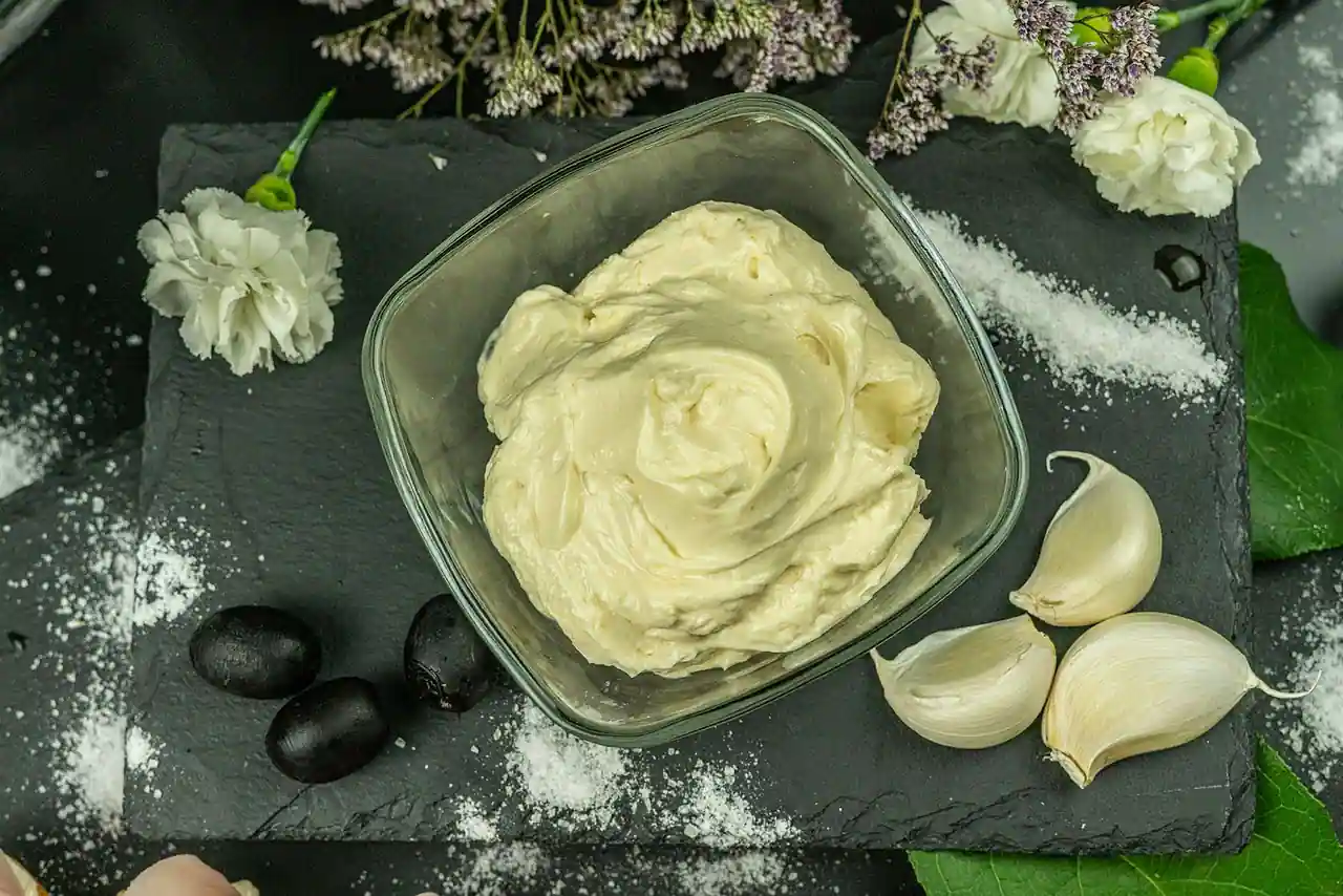 Bol en verre de mayonnaise maison crémeuse, entouré de gousses d'ail et de sel, sur une ardoise sombre.