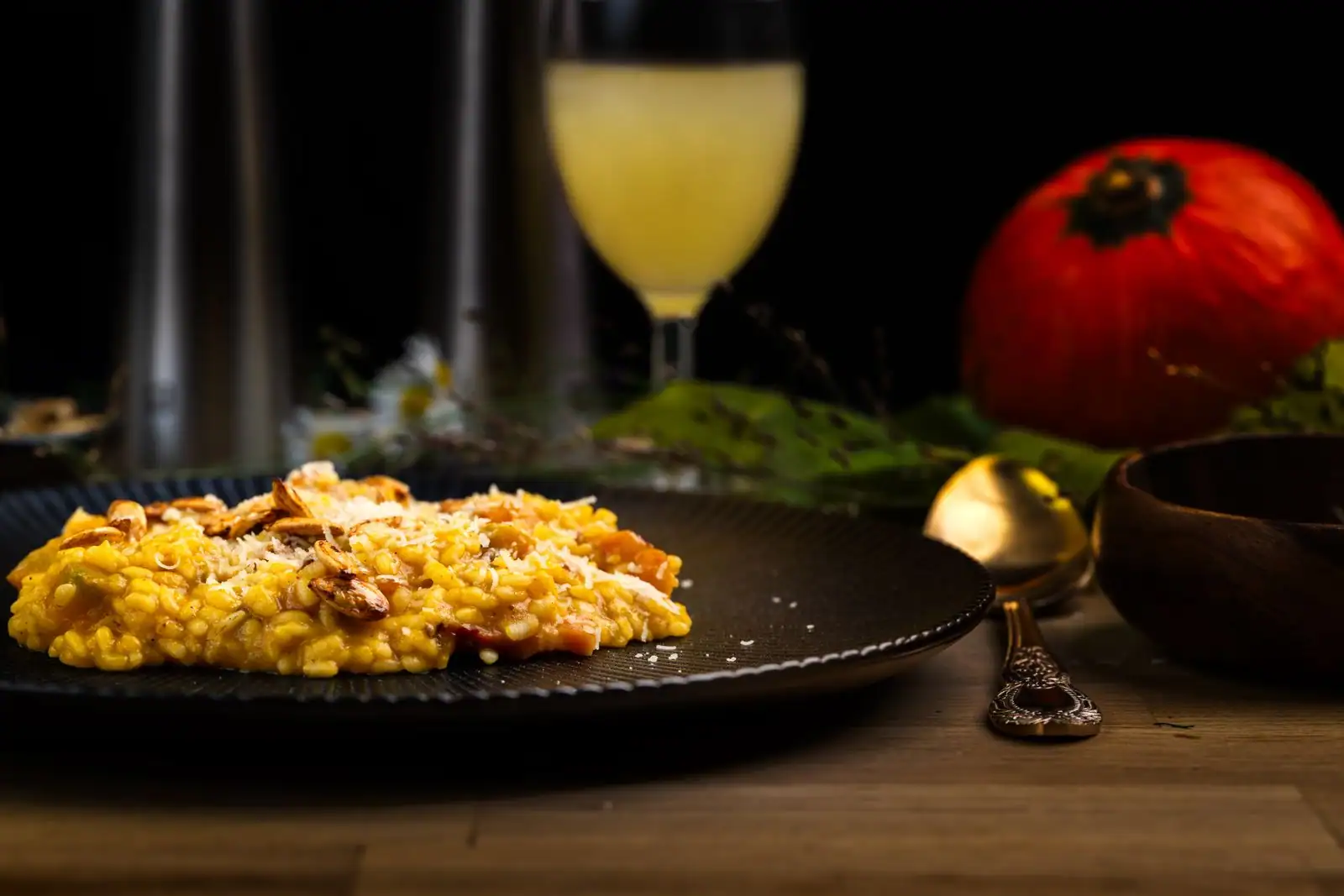 Risotto crémeux couleur safran dans une assiette sombre, parsemé de copeaux, un verre de vin blanc à l’arrière-plan.