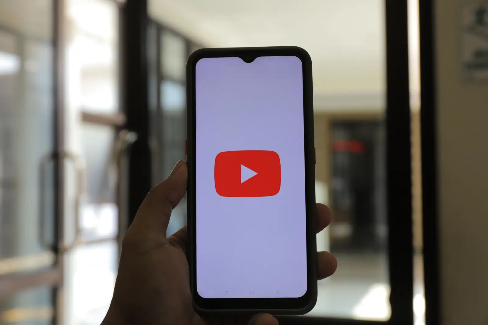 Main tenant un téléphone dont l'écran affiche le logo de YouTube, dans un couloir vitré lumineux.